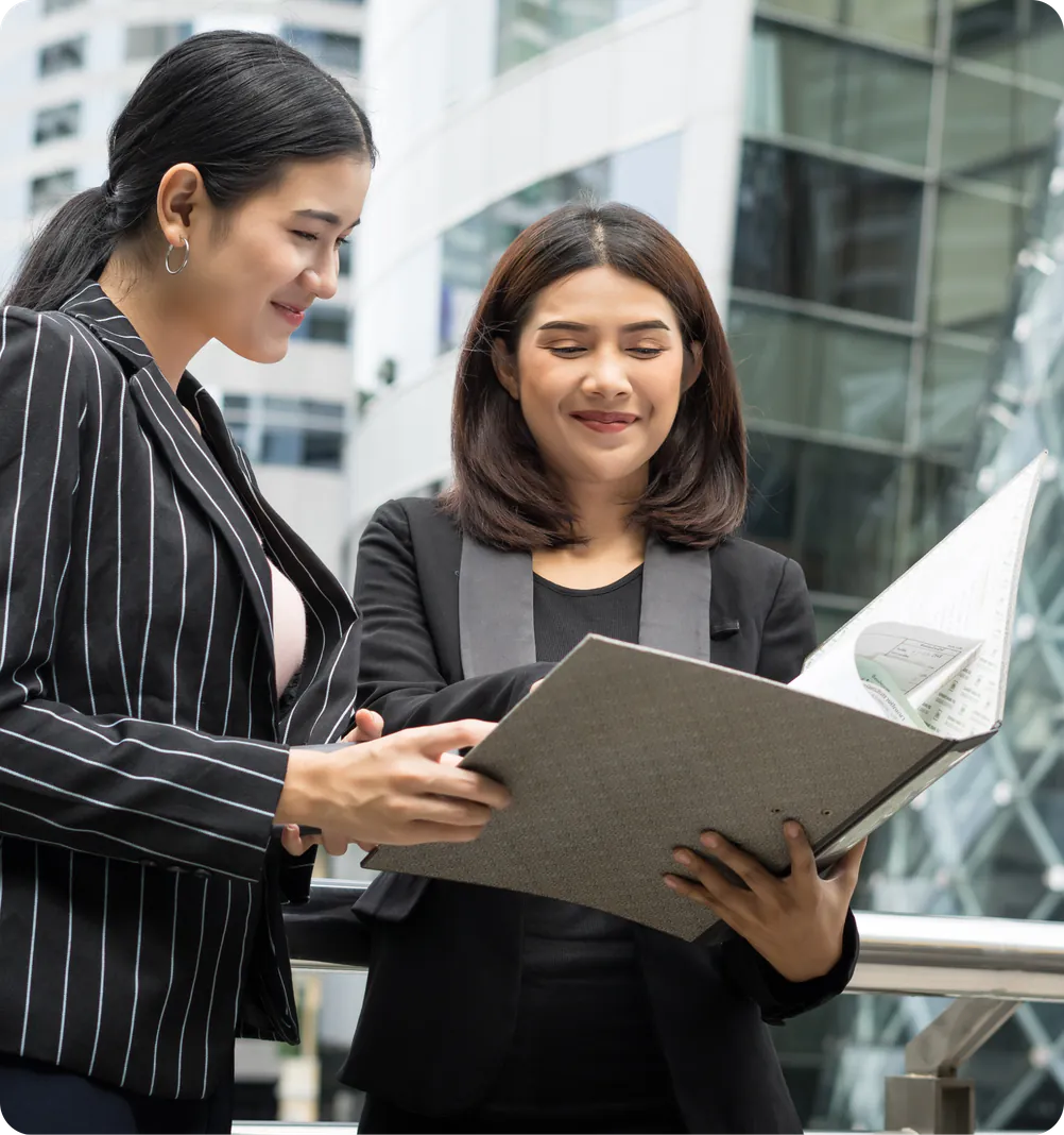 Two professional women in business attire, one in a pinstripe suit and another in a black blazer, standing outdoors near a modern office building and reviewing a document in a large folder.