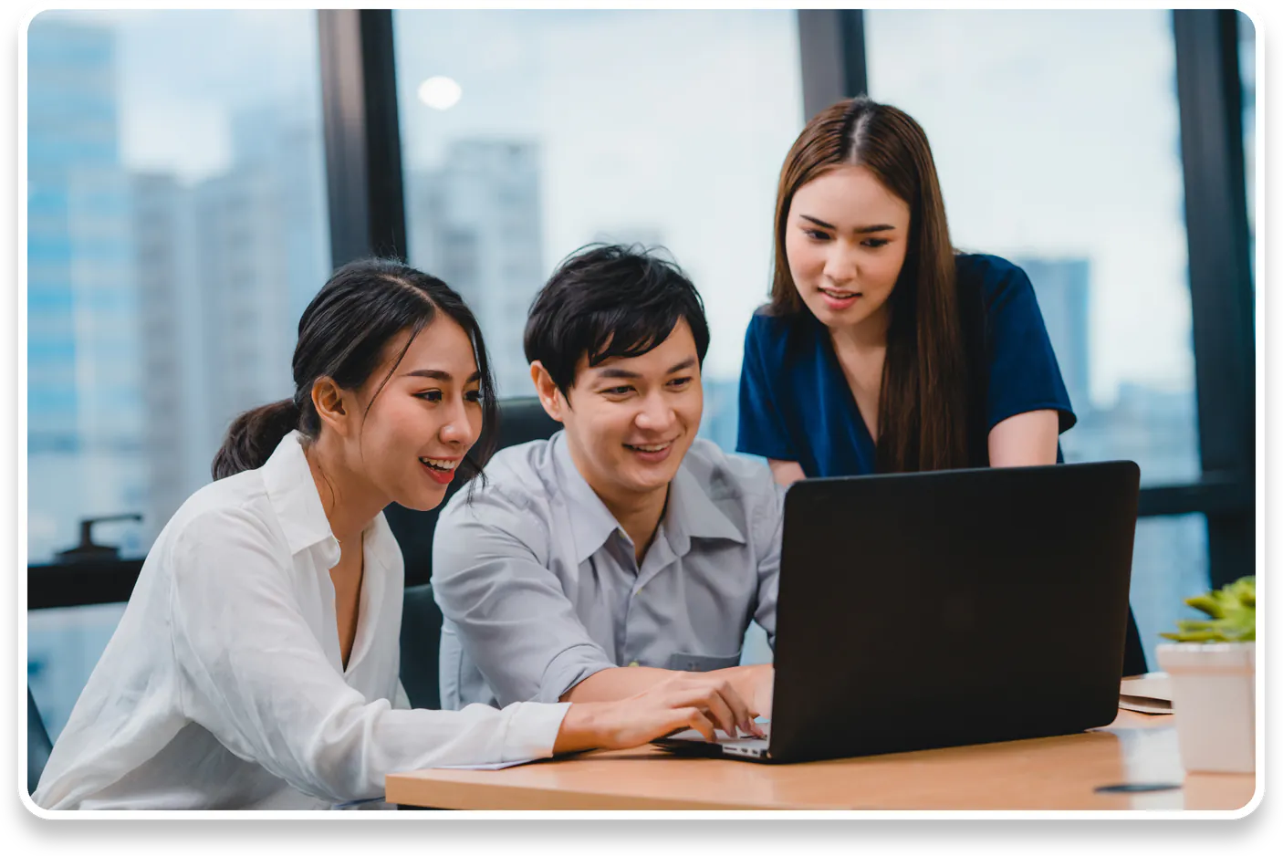 Three young professionals, two women and one man, gathered around a laptop on a wooden table in a modern office setting with large windows, looking focused and collaborative.